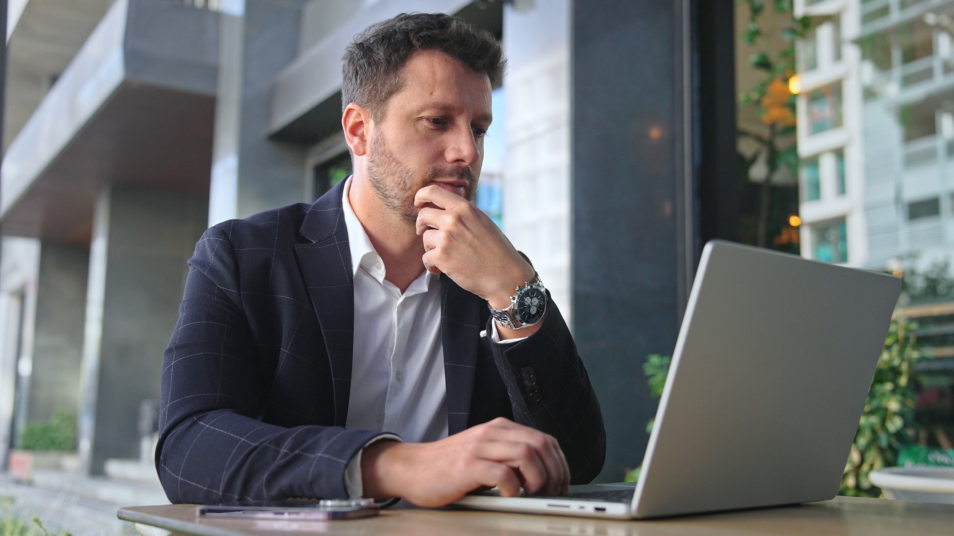 A man sits outside at a table in front of a silver laptop with his hand resting on his chin.